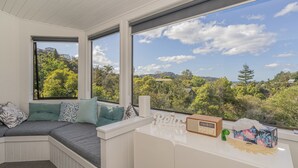 Une pièce dotée d’une banquette intégrée, de grandes fenêtres offrant une vue sur une colline boisée, et d’une petite table sur laquelle se trouvent une radio et quelques objets décoratifs.