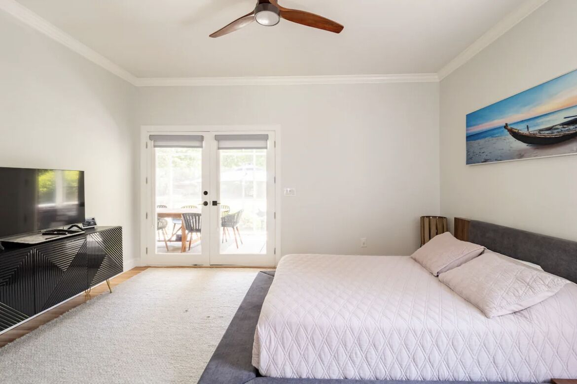 Een slaapkamer met een bed, een plafondventilator, een televisie op een zwart tv-meubel, en een schilderij van een boot aan de muur.