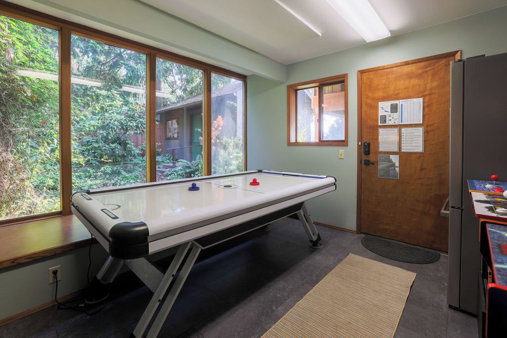 Una mesa de billar en una habitación con ventanas amplias, una puerta con papeles y un refrigerador.