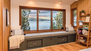 A room with a large window overlooking a lake, built-in bench seating, and a wooden bookshelf. - Private Waterfront Spa Retreat + Movie Theater (Port Townsend)