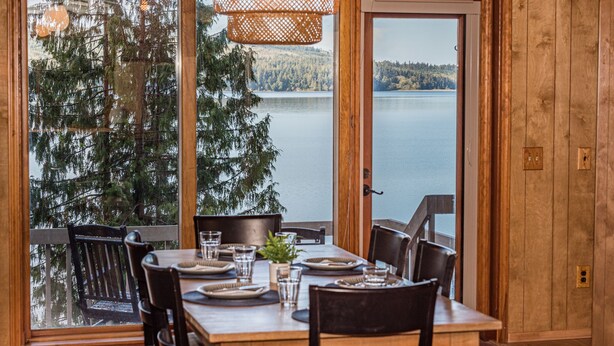 A wooden dining table set for four with a view of a lake and forest. - Private Waterfront Spa Retreat + Movie Theater (Port Townsend)