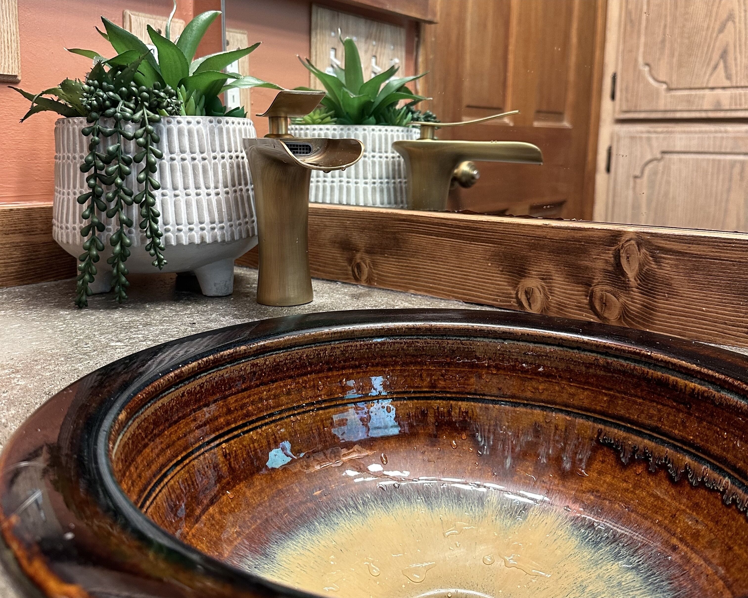 A bathroom with a large, round, brown ceramic sink, white textured vases with green plants, and a wooden door in the background.