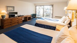Ein Hotelzimmer mit zwei Betten, einem Schreibtisch mit Computer, einem Sessel, einem Fernseher und einem Fenster mit Blick auf Gebäude.