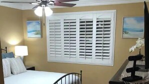 Ein Schlafzimmer mit einem Bett, Deckenventilator, einem Fenster mit Fensterläden und einem Schminktisch.