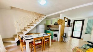 A spacious living area with a dining table and chairs, a staircase with decorative railings, a kitchen with green cabinets, and a large window with curtains. - I Geranei 2 San Teodoro Centro (San Teodoro)