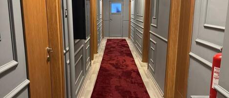 Un pasillo con alfombra roja, paredes grises, puertas de madera y un extintor.