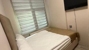 A small, single-bed room with a window, a flat-screen TV mounted on the wall, and a bedside table.