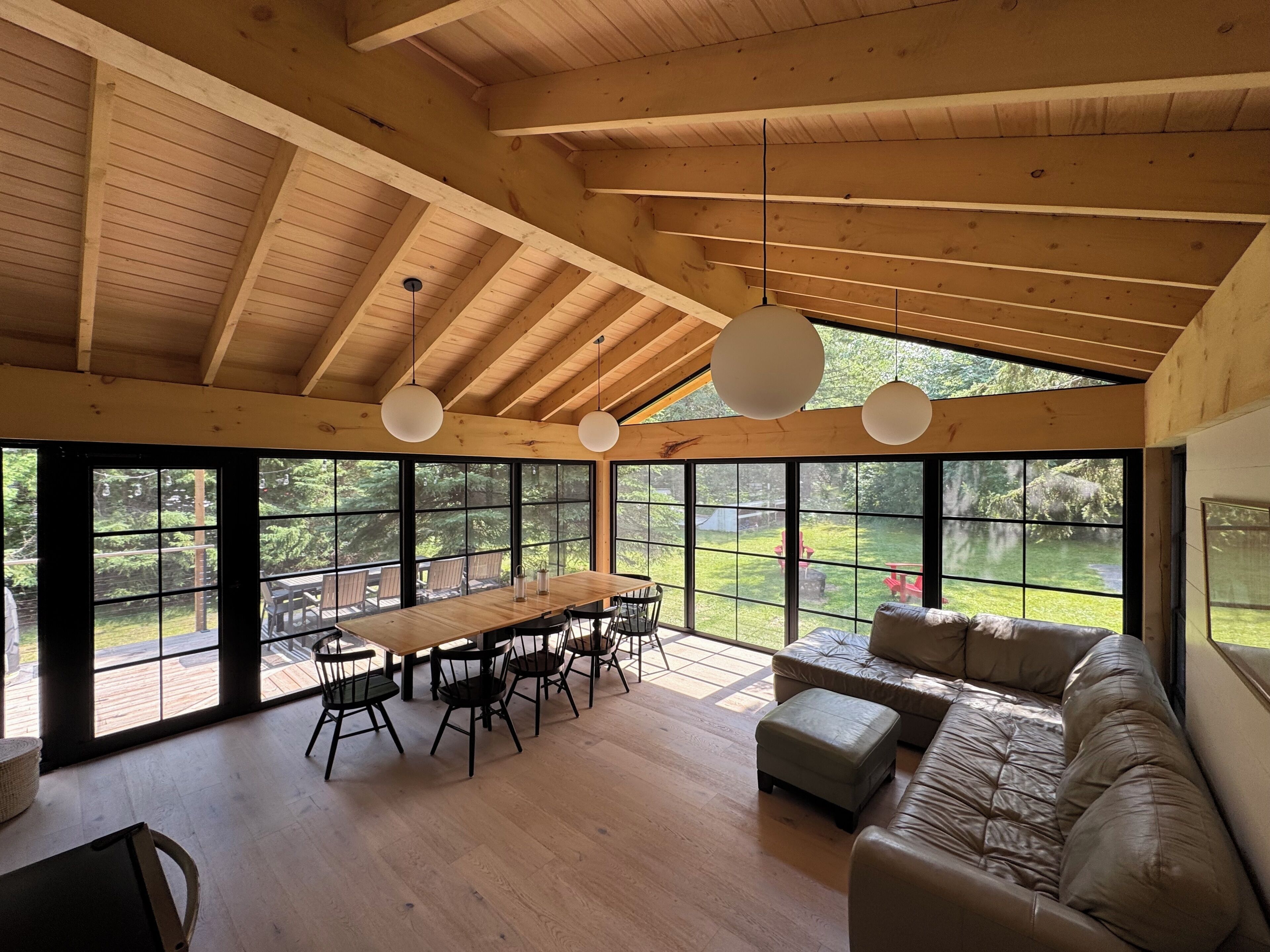 A spacious room with a high ceiling, large windows, a wooden dining table with chairs, a leather sofa, and hanging pendant lights.