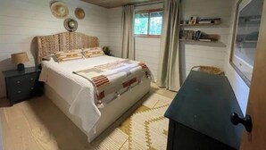 A bedroom with a wooden ceiling, a bed with patterned bedding, a nightstand with a lamp, a window with curtains, and a shelf with books.