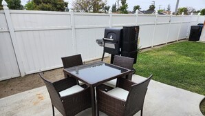 Un patio con una mesa de vidrio, cuatro sillas de mimbre, una parrilla y una cerca blanca al fondo.