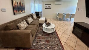 Una sala de estar con sofá, mesa de centro y alfombra estampada. Se observa un ventilador de techo, chimenea y un comedor al fondo.