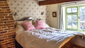 A bedroom with a brick wall, floral wallpaper, a wooden headboard, and a window with a view of the outside.