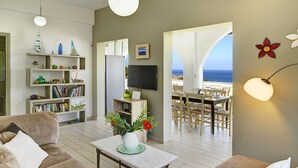 A modern living room with a sofa, coffee table, and TV. There are shelves with books and decorative items, and a dining area with a table and chairs.