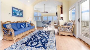 A spacious living room with a blue sofa, a patterned rug, and wicker furniture. - Oceanfront Home with Hot Tub, Pool, Putting Green, Elevator, and Game Room (Avon)