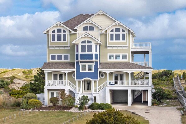 A multi-story house with a prominent front porch, a balcony on the second floor, and a garage. - Oceanfront Home with Hot Tub, Pool, Putting Green, Elevator, and Game Room (Avon)
