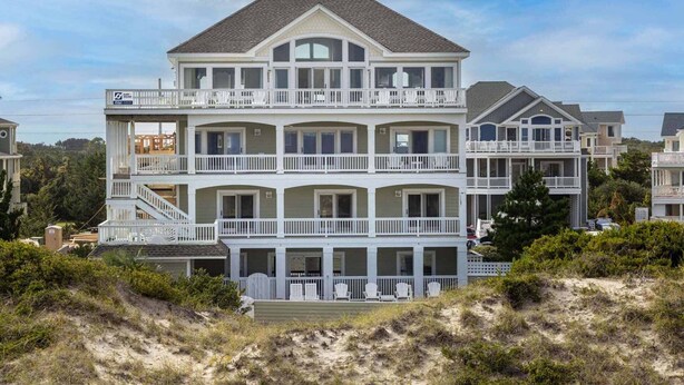 A multi-story beachfront house with balconies, a covered porch, and a sandy landscape. - Oceanfront Home with Hot Tub, Pool, Putting Green, Elevator, and Game Room (Avon)