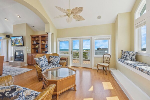 A spacious living room with a fireplace, wicker furniture, a large glass coffee table, and a view of the ocean through the sliding glass doors. - Premier Oceanfront Home w/ Pool, Hot Tub, Elevator, Putting Green, Game Room (Avon)