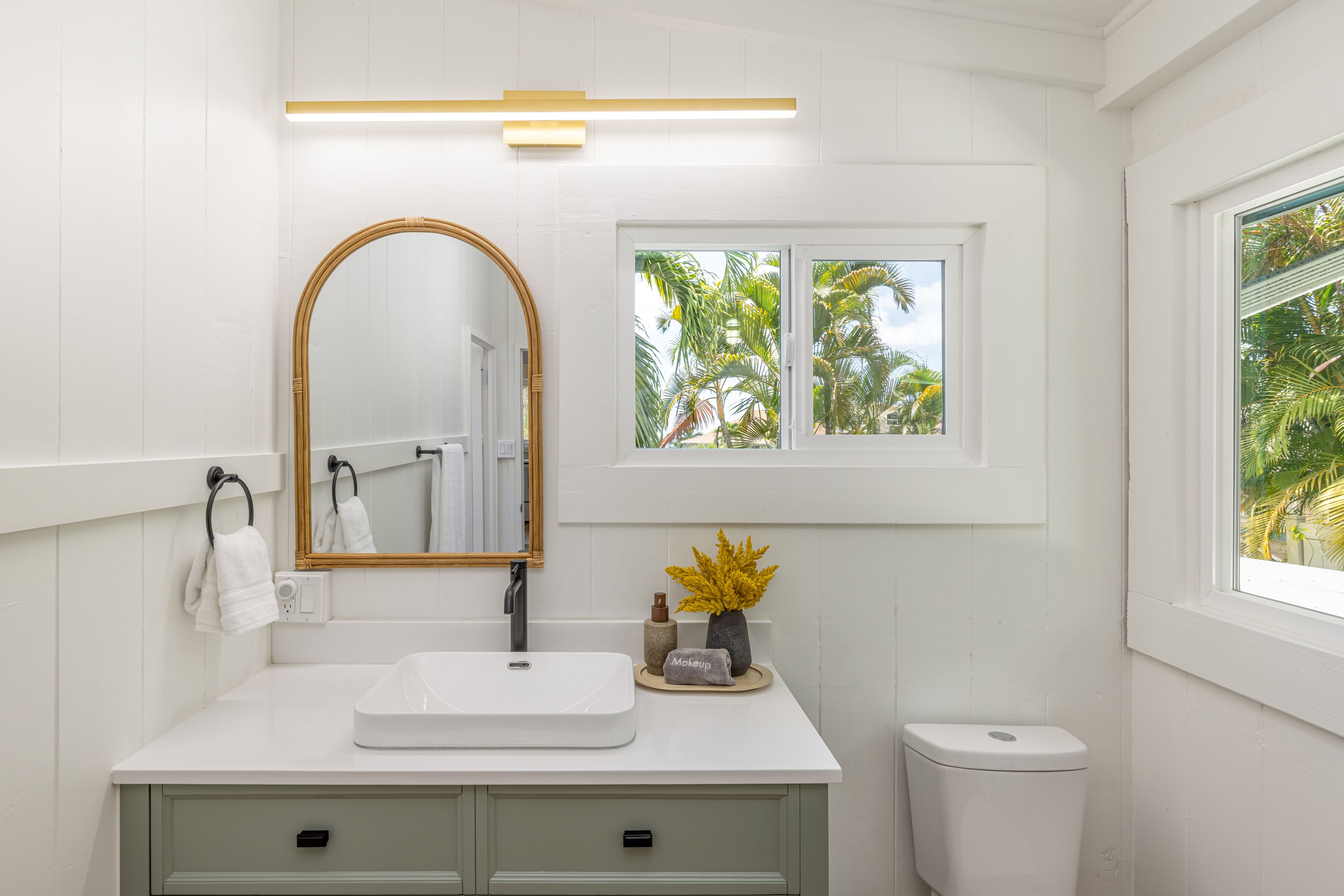 A bathroom with a white vanity, a mirror, a window with a view of palm trees, and a toilet.