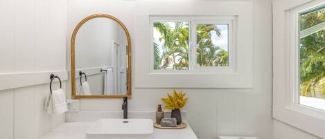 A bathroom with a white vanity, a mirror, a window with a view of palm trees, and a toilet.