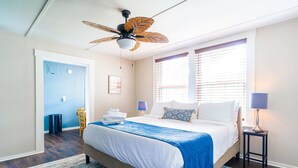 Una habitación de hotel con una cama, un ventilador de techo, una ventana con persianas, una mesita de noche y una lámpara.