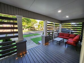 Eine überdachte Terrasse mit rotem Sofa, einem kleinen Tisch und Blick auf eine Straße sowie Grünflächen.
