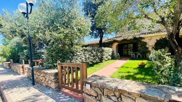 Un sendero de piedra que conduce a un edificio con techo de tejas, rodeado de abundante vegetación y un portón de madera.