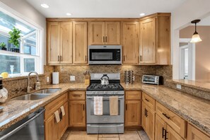 A modern kitchen with wooden cabinets, granite countertops, stainless steel appliances, and a window with plants. - Beach Home-Rooftop Deck-Steps to Sand (Oxnard)