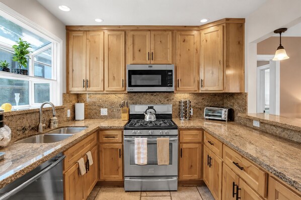 A modern kitchen with wooden cabinets, granite countertops, stainless steel appliances, and a window with plants. - Beach Home-Rooftop Deck-Steps to Sand (Oxnard)