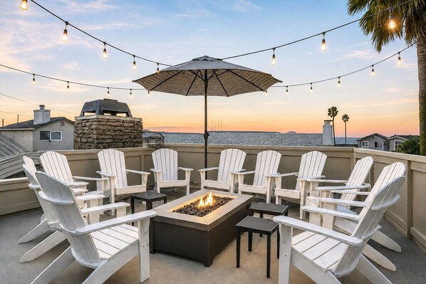A rooftop patio with a fire pit, white Adirondack chairs, string lights, and a view of the ocean at sunset. - Spacious Beach Home-Rooftop Deck-Steps to Sand (Oxnard)
