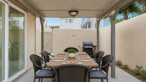 Une terrasse extérieure avec une table dressée pour quatre personnes, entourée de chaises en osier, d’un barbecue et d’une plante en pot.