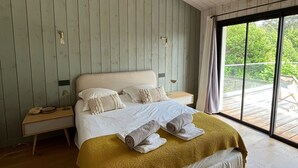 Une chambre avec un plafond en bois, un grand lit avec une literie blanche, un plaid jaune et une table de chevet avec une lampe.