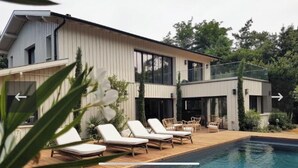 Une maison moderne dotée d’une piscine, de chaises longues et d’un patio fermé par des baies vitrées.
