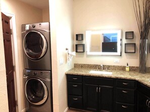 A laundry area with two stacked front-loading washers and dryers, a granite countertop with a sink, and a mirror above the countertop.