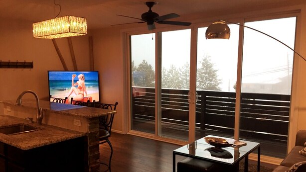 A modern living room with a kitchen area, a large TV, and a balcony with a view of the outdoors. - Lookout Mountain Lodge & Chalet with Views! (30 Day Min) (Golden)