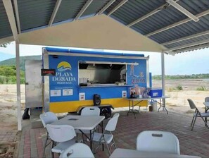 Un camión de comida con exterior azul y amarillo, identificado con las leyendas "Playa Dorada Beach Club" y "El Rincón". Está estacionado bajo un área techada con mesas y sillas blancas.