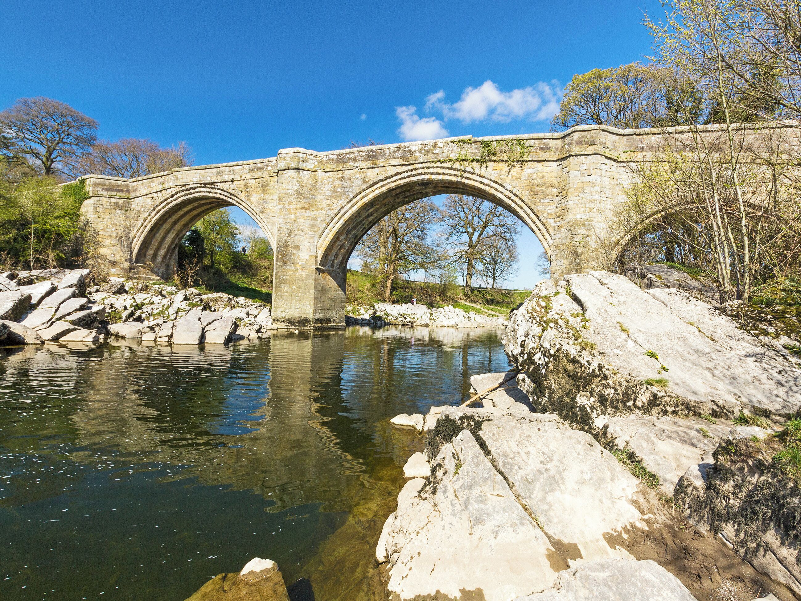 A stone bridge over a river with clear water, surrounded by rocks and trees.