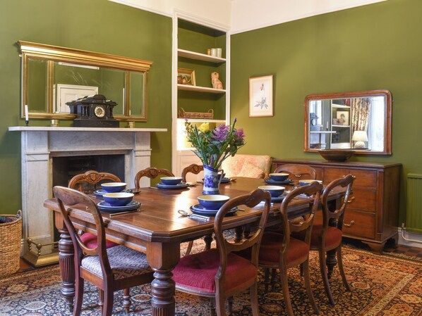 A dining room with a wooden table set for six, surrounded by chairs. There is a fireplace with a mantel, and a bookshelf to the right.
