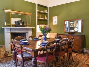 A dining room with a wooden table set for six, surrounded by chairs. There is a fireplace with a mantel, and a bookshelf to the right.