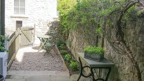 Un sendero de piedra conduce a una zona de estar con una mesa y sillas, rodeada de vegetación y un muro de piedra.