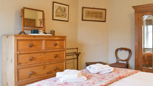 Un dormitorio con un aparador de madera, una cama con colcha estampada, una silla y dos cuadros en la pared.