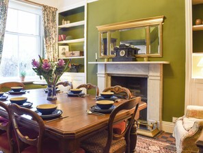 A dining room with a wooden table set for four, surrounded by chairs. A fireplace is visible in the background, and there is a mirror above it.