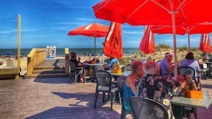 Een restaurant aan het strand met terras, rode parasols en uitzicht op de oceaan.