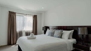 A hotel room with a large bed, a nightstand, a lamp, and a window with curtains. - Urban 1-Bed Apartment in the Heart of the City (Braddon)