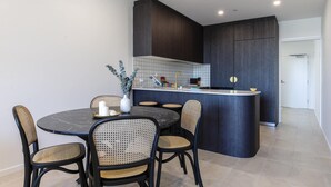 Une cuisine moderne avec des meubles de couleur sombre, un plan de travail blanc et un coin repas doté de chaises en osier et d’une table en marbre.