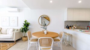 Ein modernes Wohnzimmer mit einem runden Holzesstisch, weißen Korbstühlen, einem weißen Sofa und einer Küchenzeile mit weißer Arbeitsplatte und sechseckiger Rückwand.