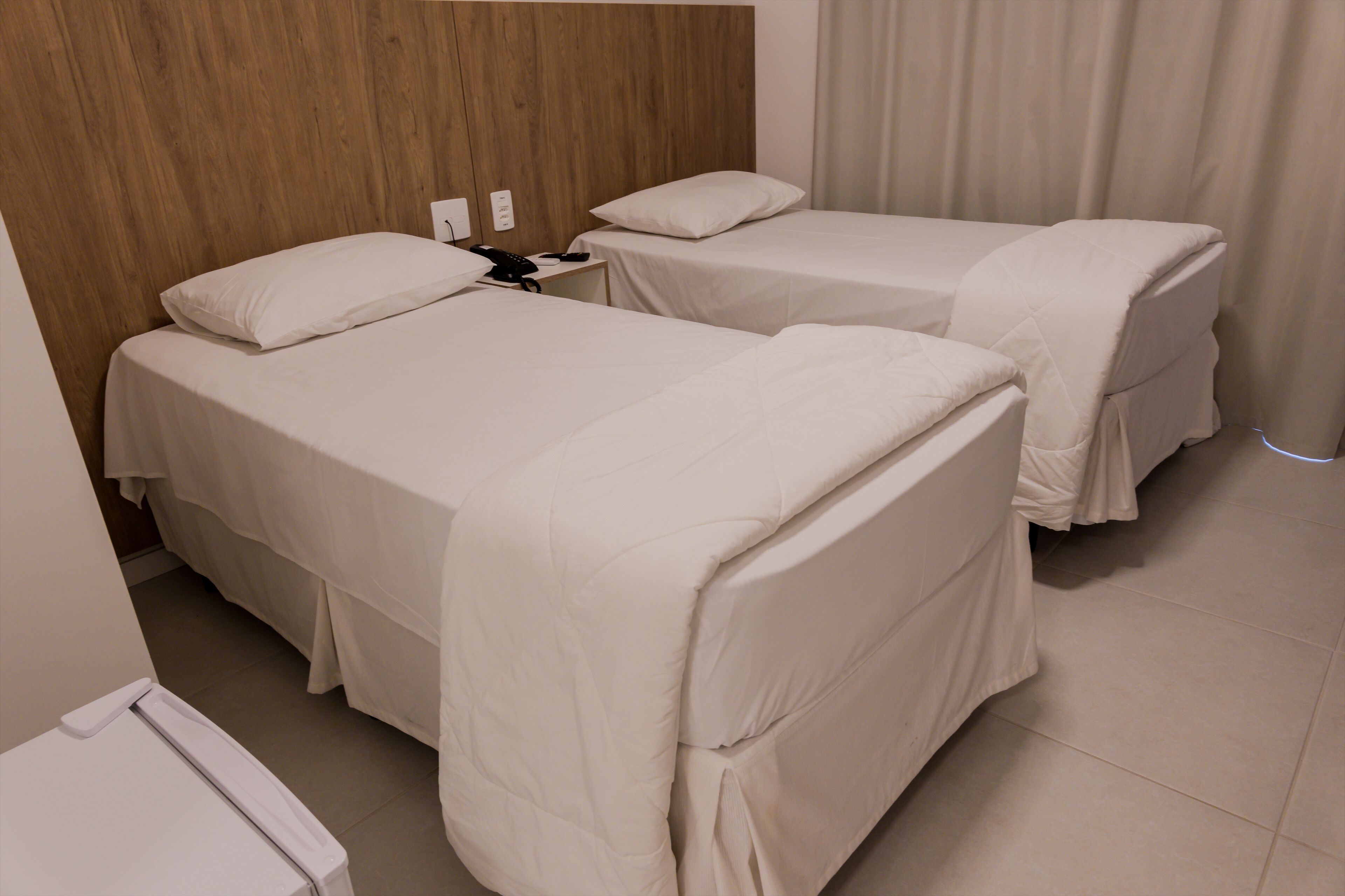 Une chambre d’hôtel avec deux lits simples, une tête de lit en bois, une table de chevet avec un téléphone et un rideau.