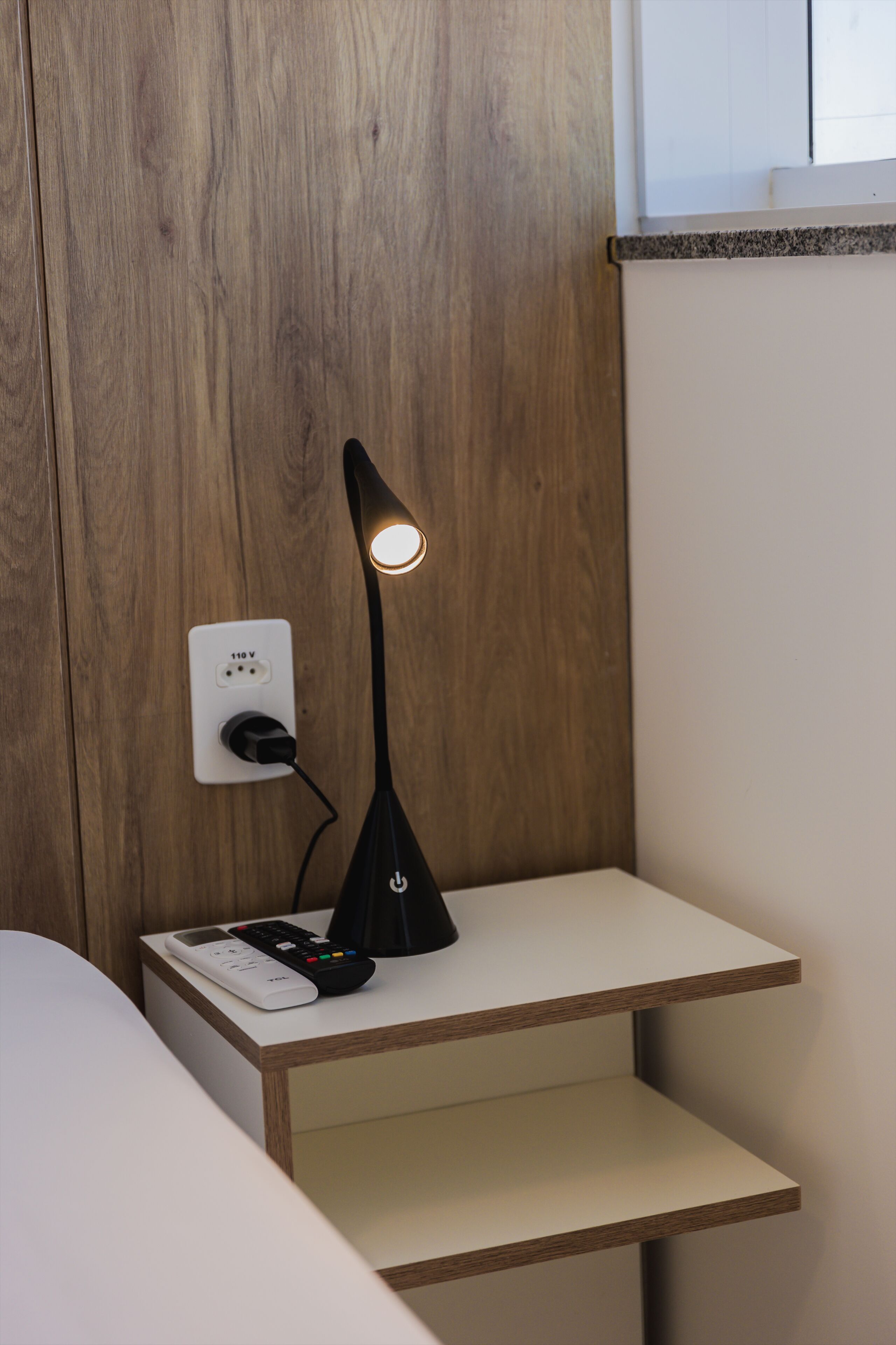 A bedside table with a lamp, a power outlet, and a remote control.