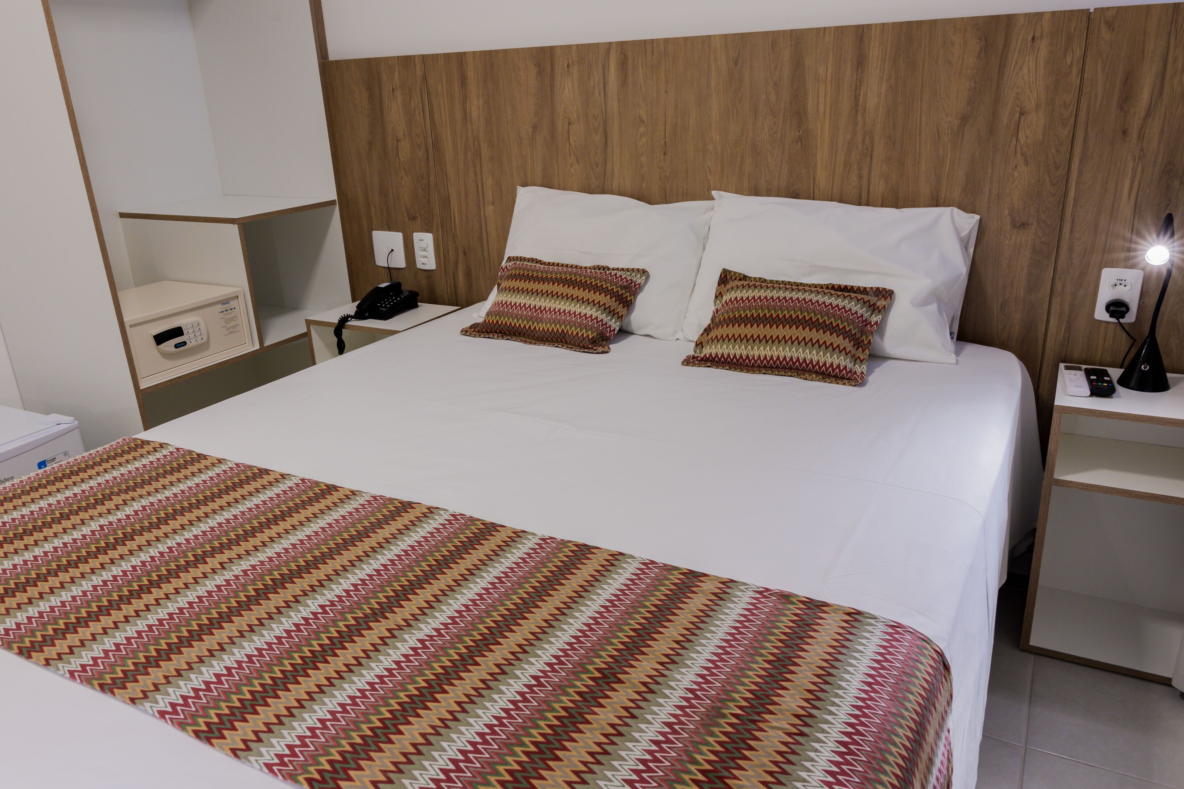 Una cama bien hecha con sábanas blancas y dos almohadones estampados, una mesita de noche con una lámpara, y un estante pequeño con un teléfono y una caja.
