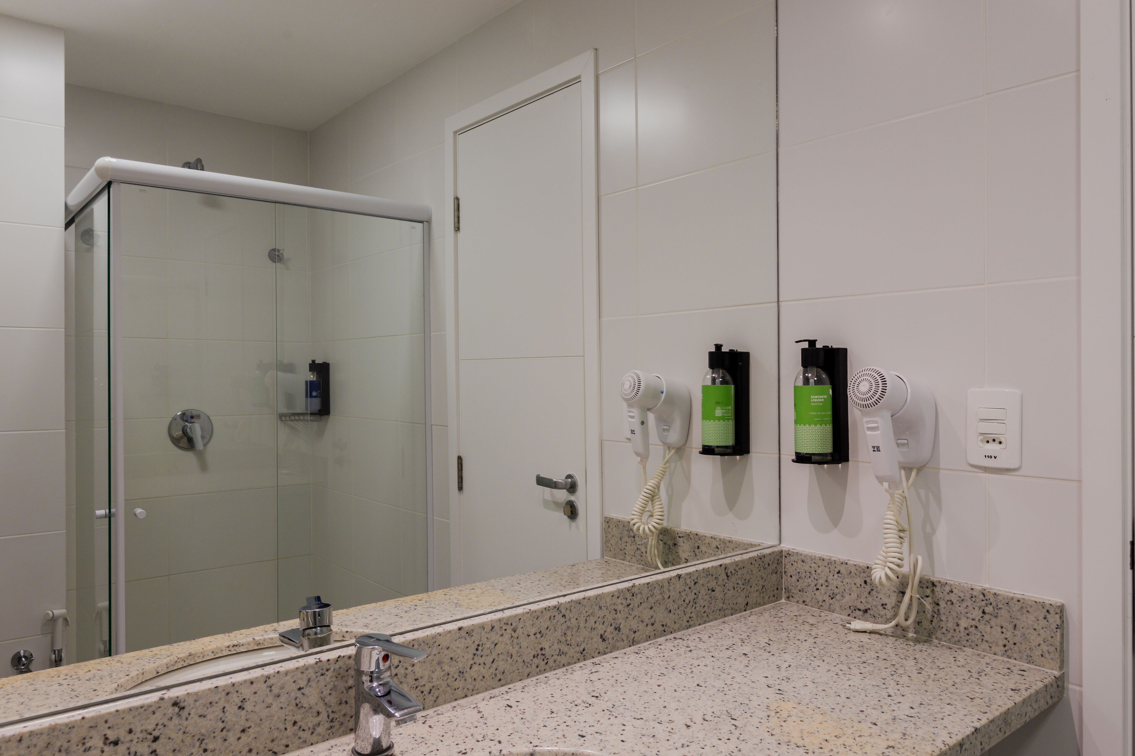 A bathroom with a glass shower enclosure, a sink with a granite countertop, and two hair dryers mounted on the wall.