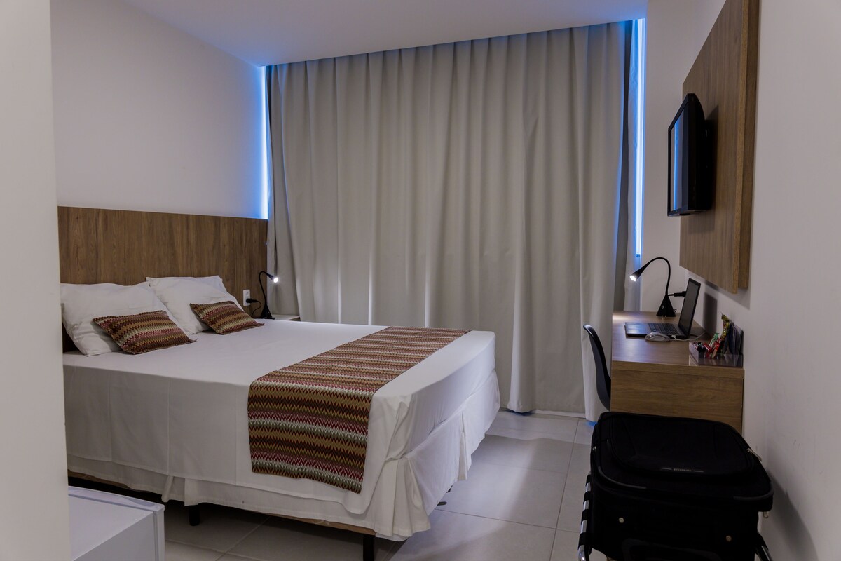 Una habitación de hotel con una cama, un escritorio con una computadora, un televisor y una maleta.